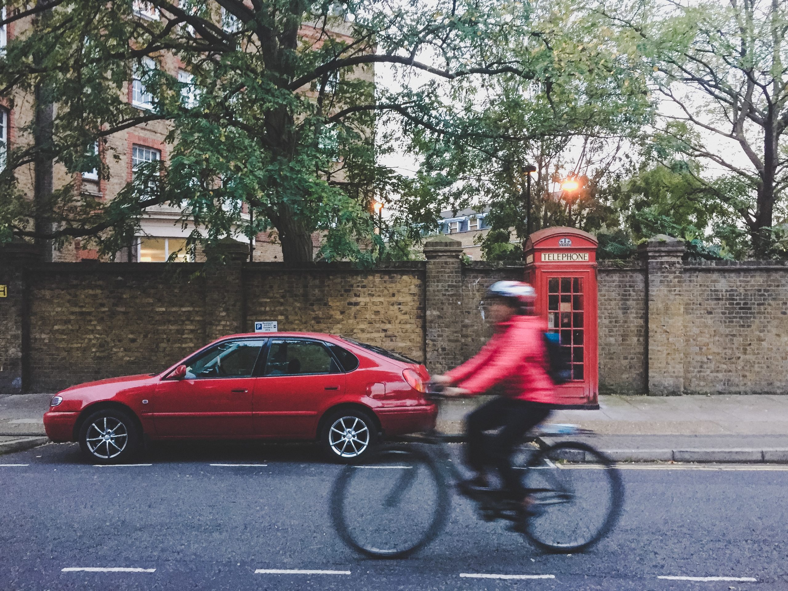A cyclist in motion on the road, with a parked car and telephone box in the background