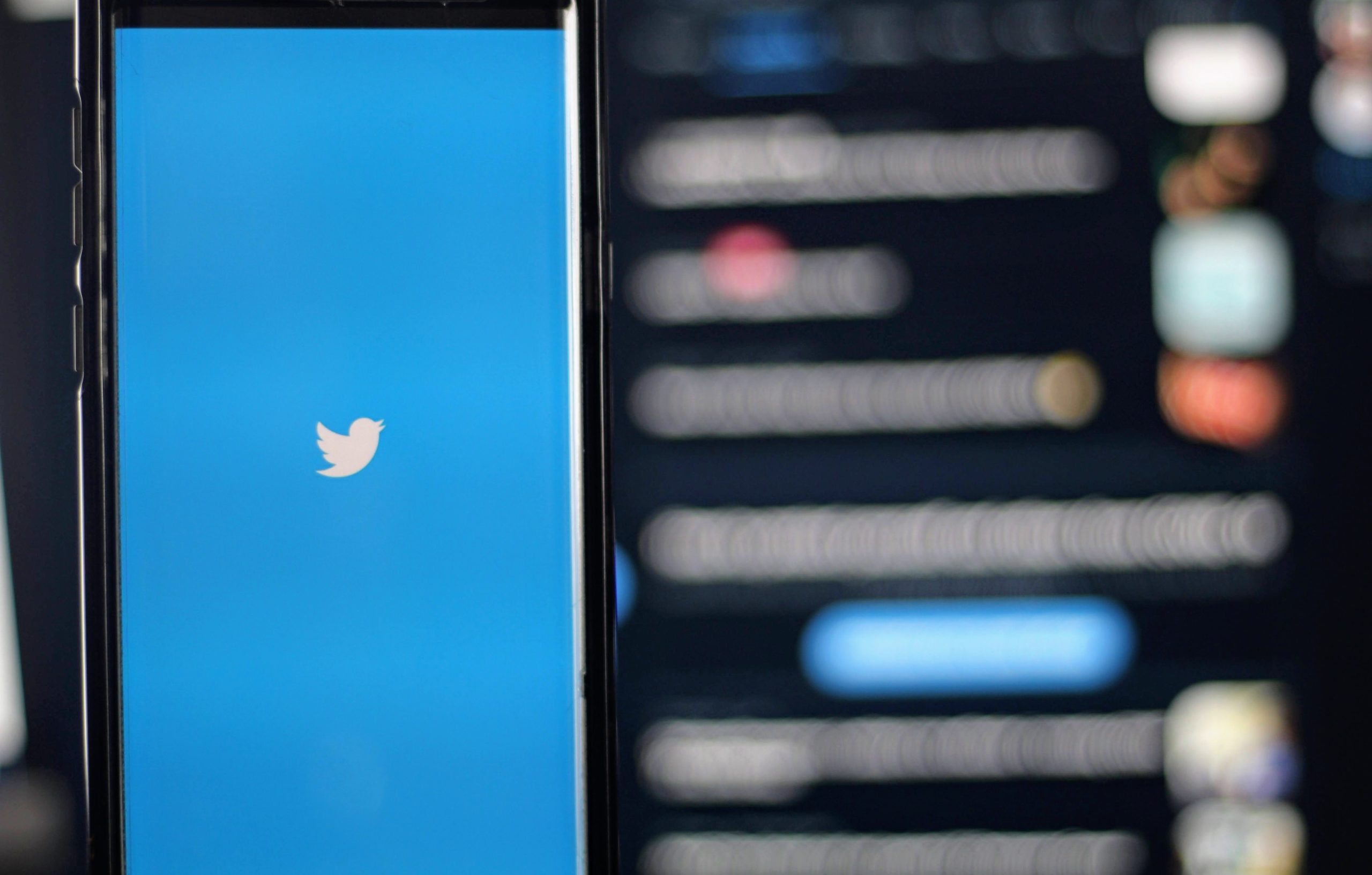 Smartphone displaying the Twitter icon (white bird on blue background), with a blurred laptop screen behind