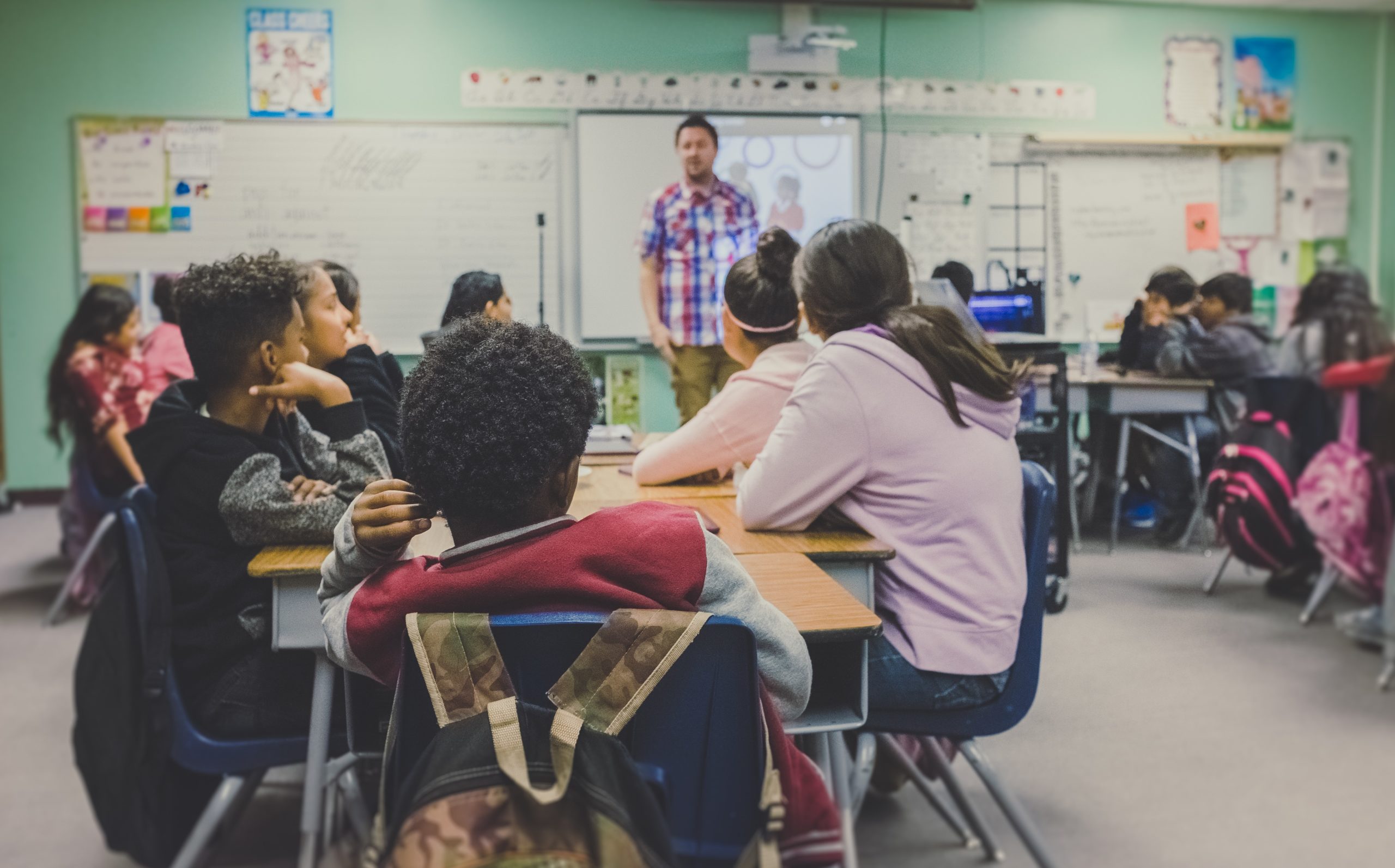 A school classroom, with students sitting around desks, looking at the teacher standing at the front of the classroom by the whiteboard