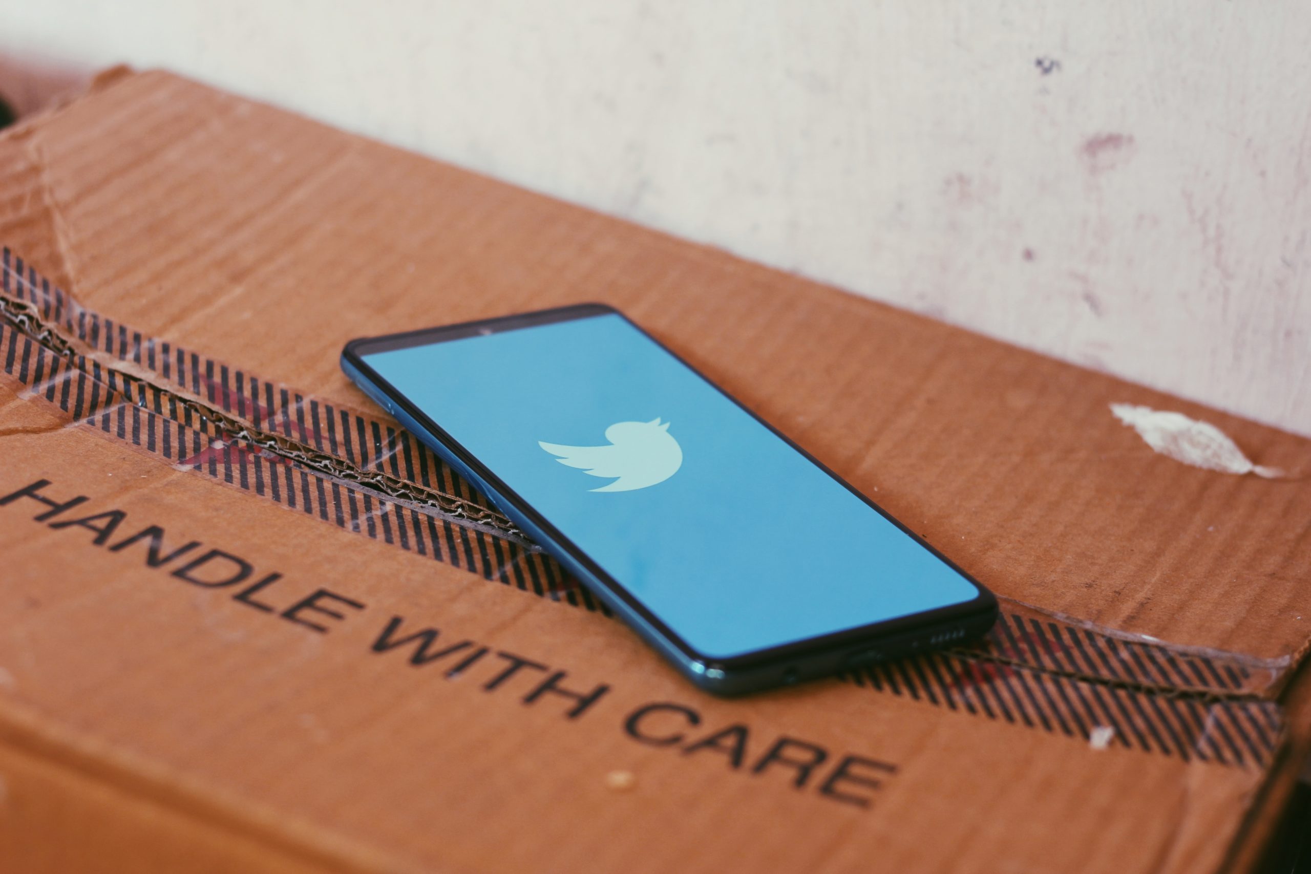 A smartphone with the Twitter logo onscreen, lying on a cardboard box with the words 'Handle with care' on it.