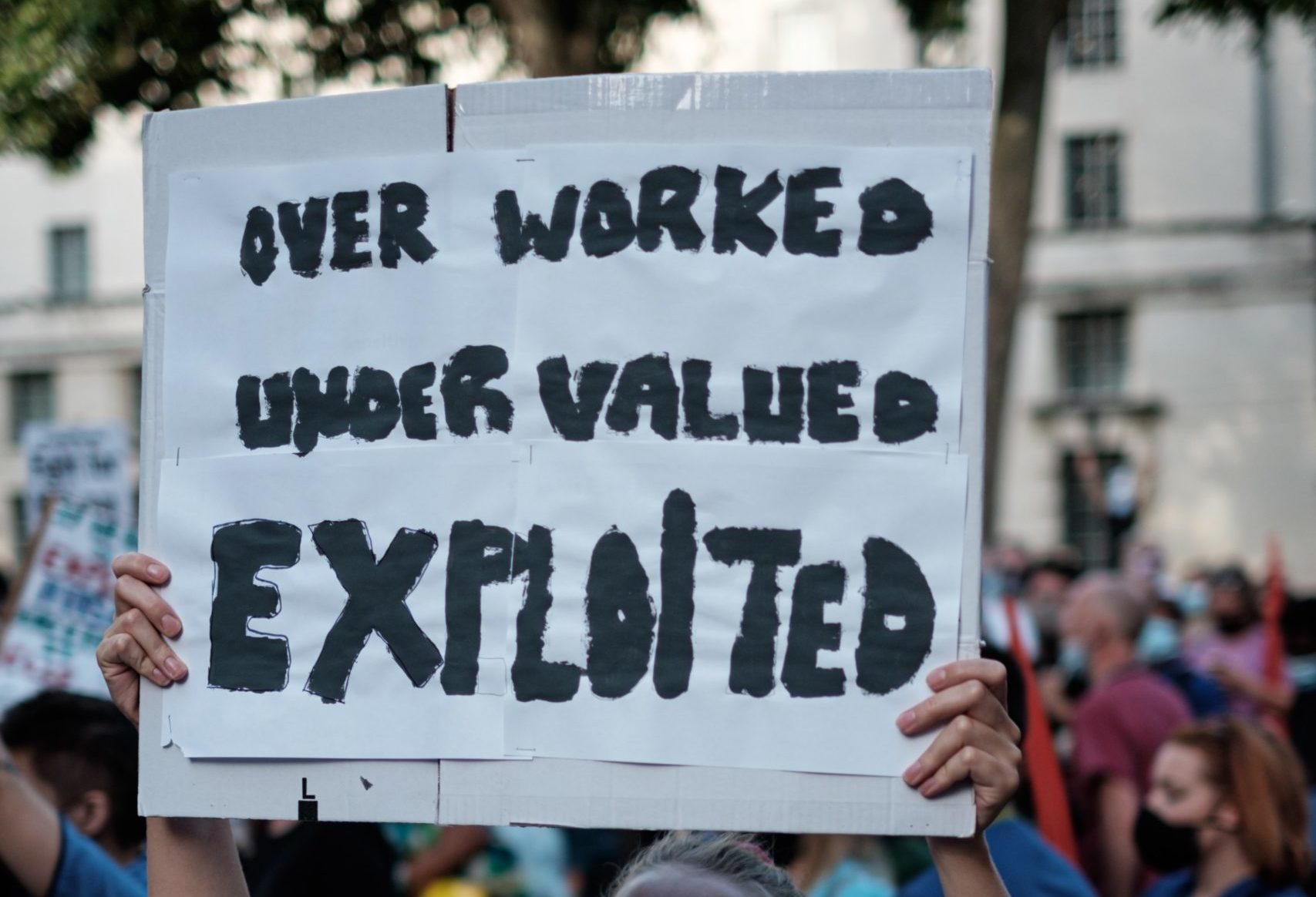 A woman with a mask on holding up a sign saying Over Worked, Under Valued, Exploited