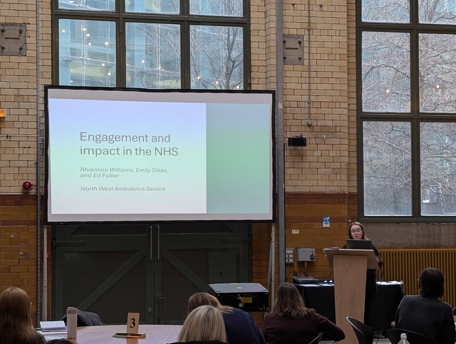 Rhiannon Williams speaking a a podium in front of a presentation titled "Engagement and Impact in the NHS"