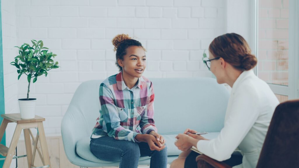 Photo of two people sitting on chairs opposite each other. One has a notebook and pen and is facing away from the camera, the other is talking with their hands closed in front of them. There is a plant on a stool to the left of the people.
