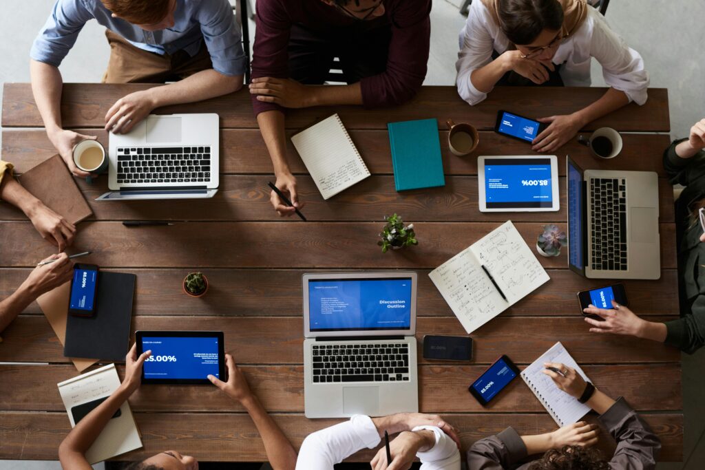 Birds eye view of a brown wooden table with eight people sitting around it. The people have a mix of laptops, cups, notebooks, pens and tablets in front of them. 